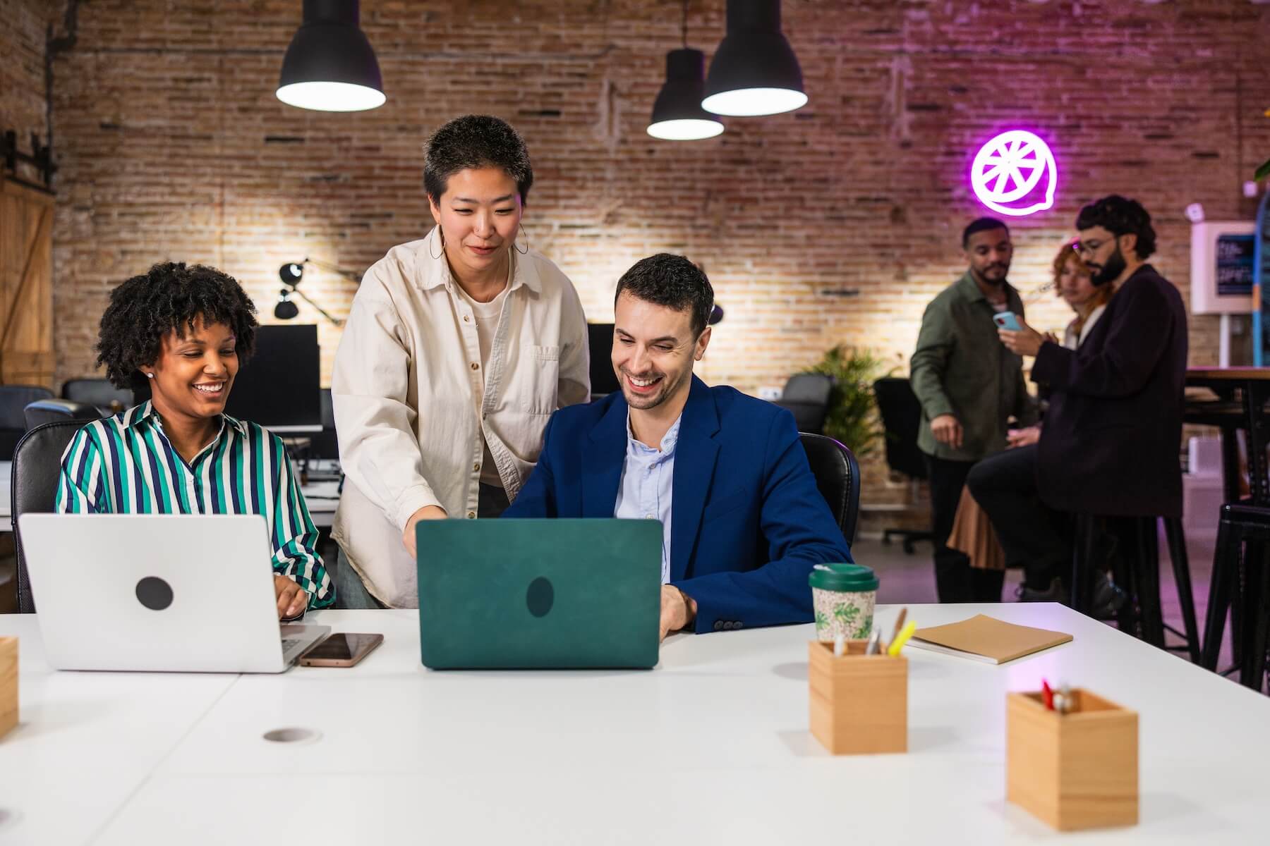 IMG-DVZH324.jpg Diverse team collaborating on laptops at a modern office workspace with exposed brick and neon sign.