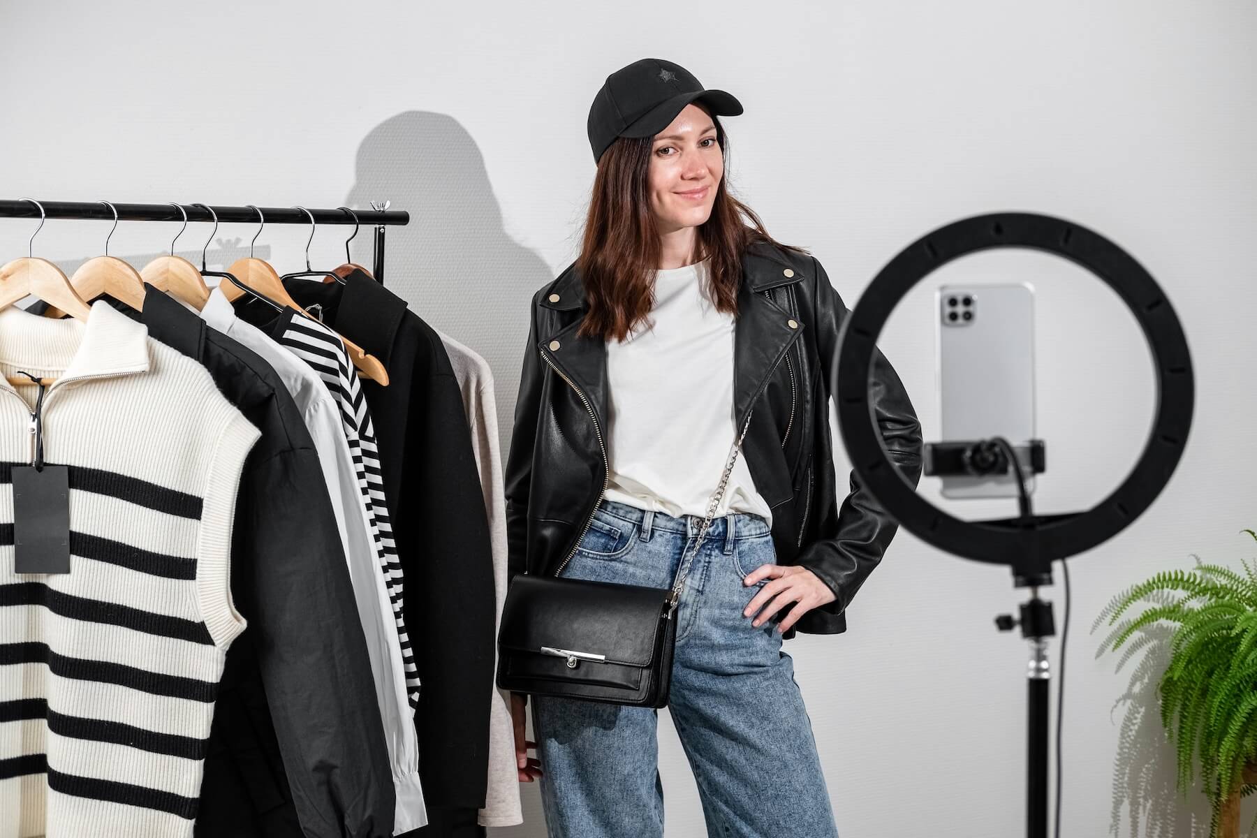 Woman in jeans and leather jacket with clothing rack and ring light.